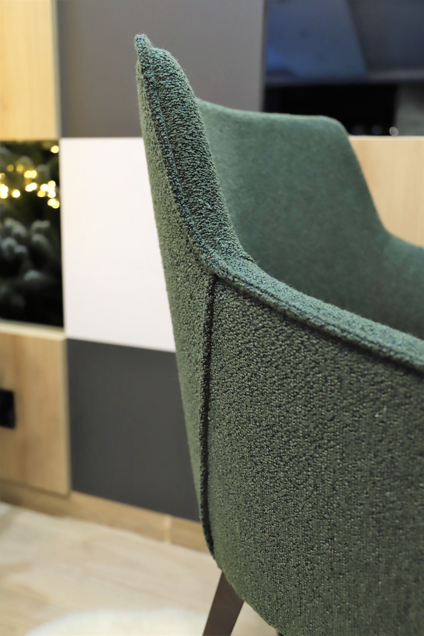 Fragment zielonego fotela z tapicerką o wyraźnej fakturze, widoczna część oparcia i siedziska, na tle ściany w geometryczne wzory i choinki z lampkami.