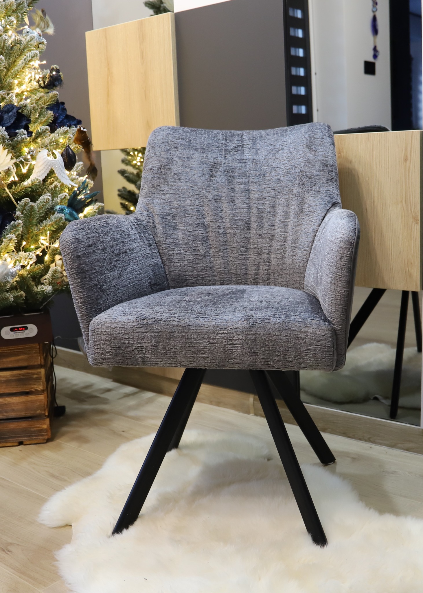 Tapicerowane krzesło w odcieniach szarości na czarnych nogach, ustawione na białym, puszystym dywanie, z choinką w tle.