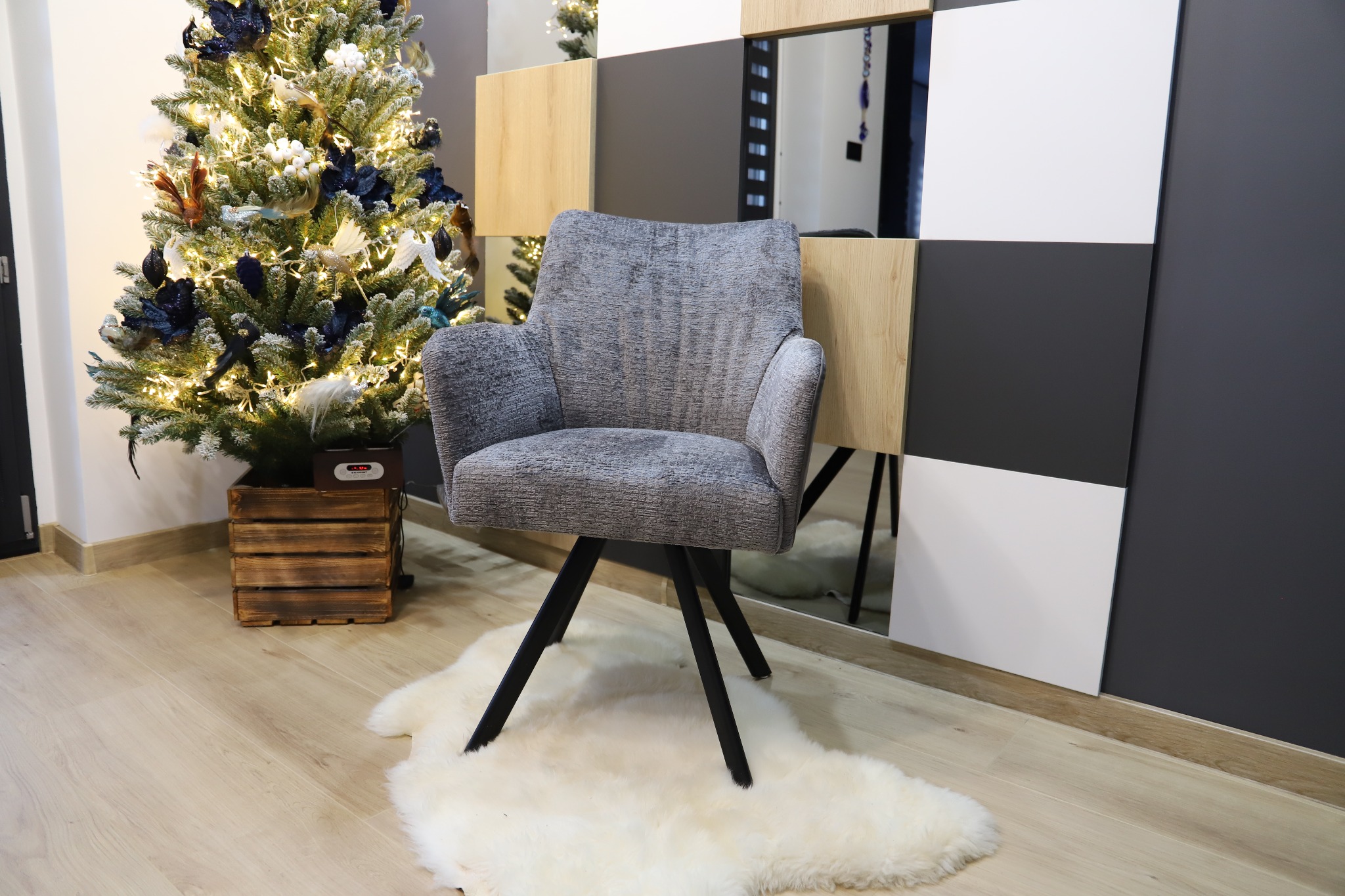 Szary fotel tapicerowany na czarnych nogach, ustawiony na białym dywanie ze skóry owczej, obok choinki w drewnianej skrzyni, na tle ściany z kwadratowymi panelami w kolorach drewna, bieli i szarości.