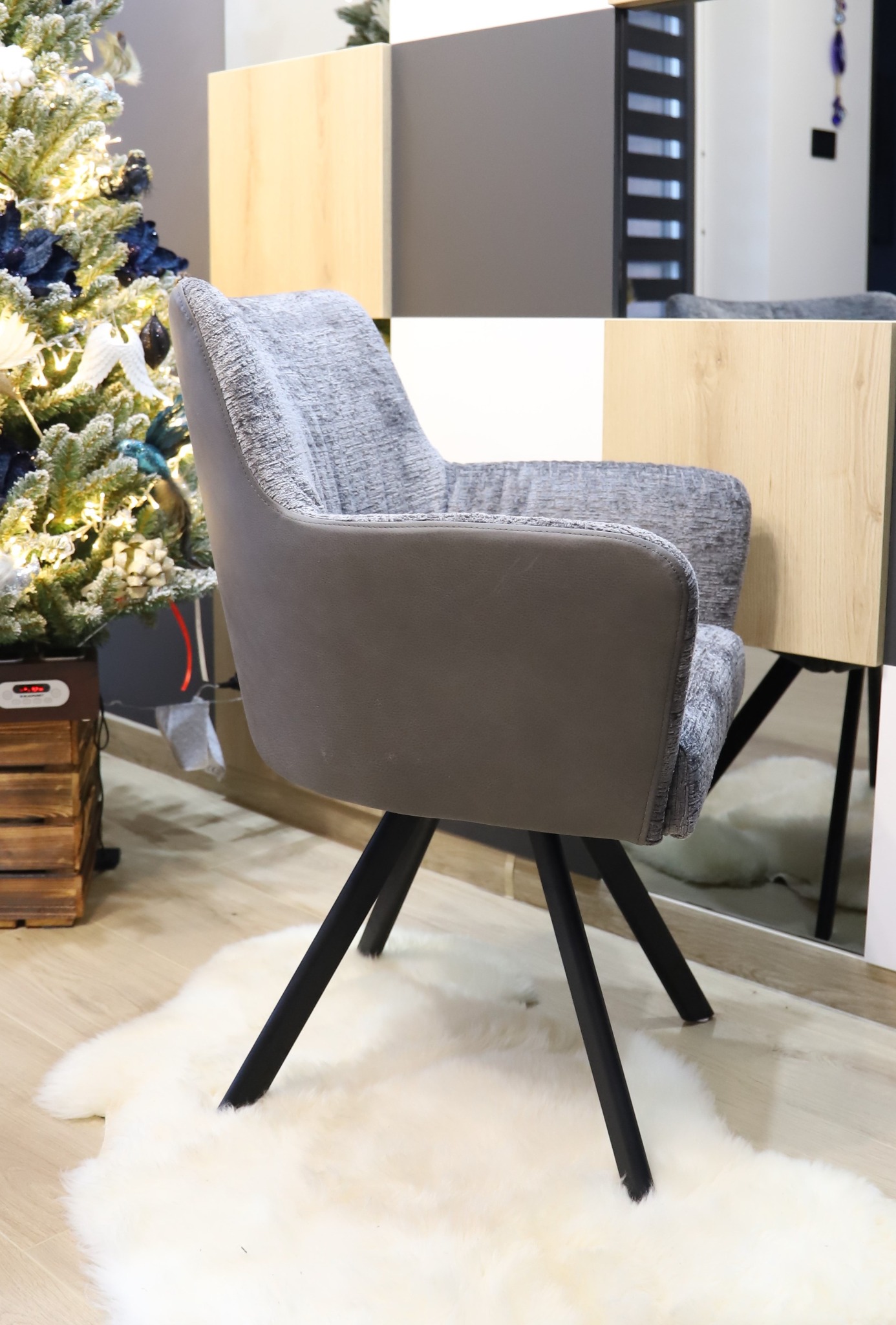 Szare krzesło z podłokietnikami na czarnych nogach, ustawione na białym futrzanym dywaniku, obok choinki i mebli z elementami drewna.