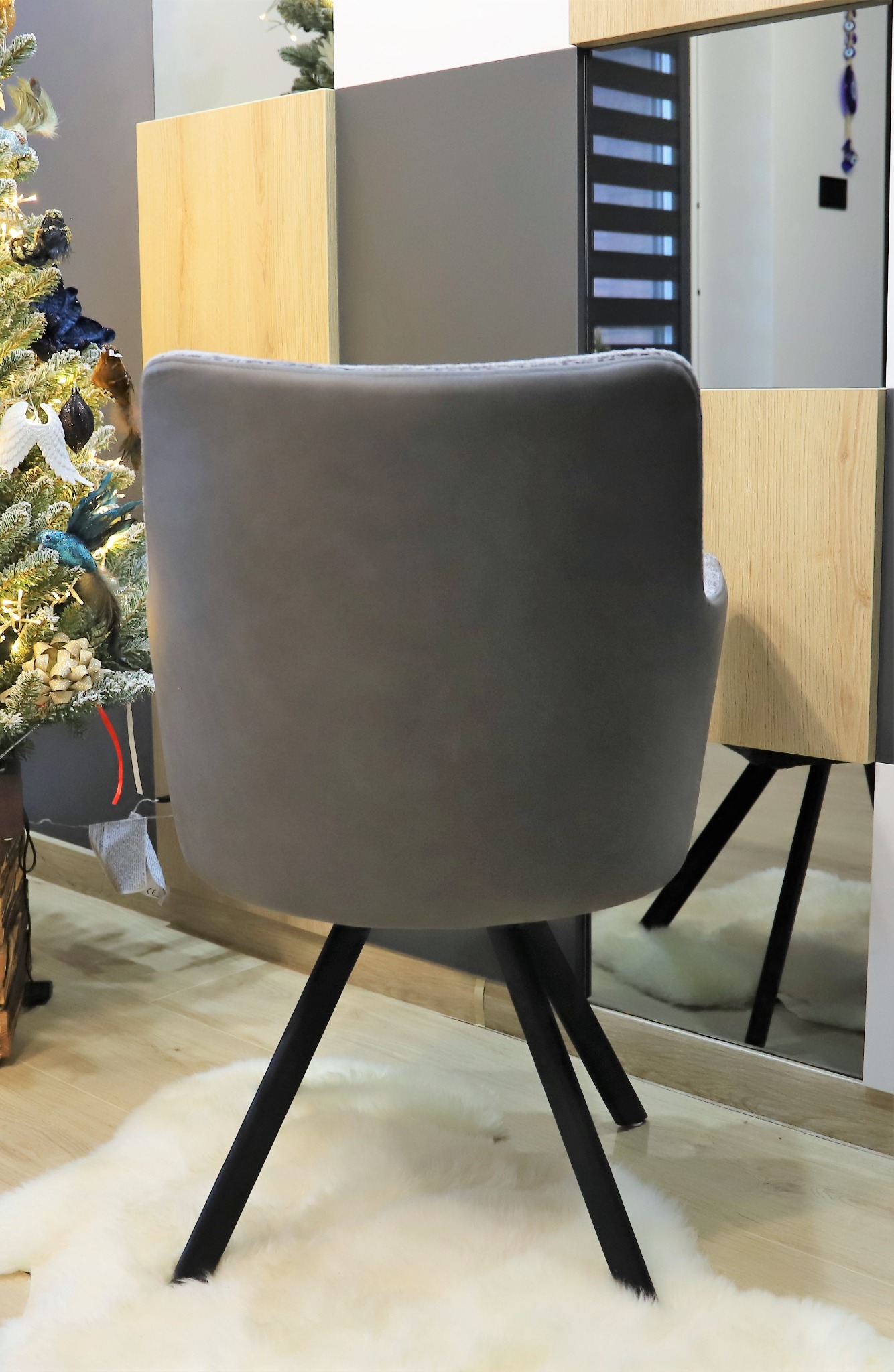 Szare krzesło tapicerowane z czarnymi nogami na tle lustra i choinki, widok z tyłu.