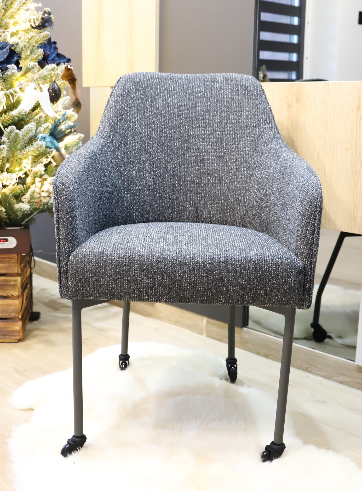 Szare tapicerowane krzesło z podłokietnikami i kółkami na nóżkach, umieszczone na białym dywaniku z długim włosiem, w tle choinka i biurko z drewnianym blatem.