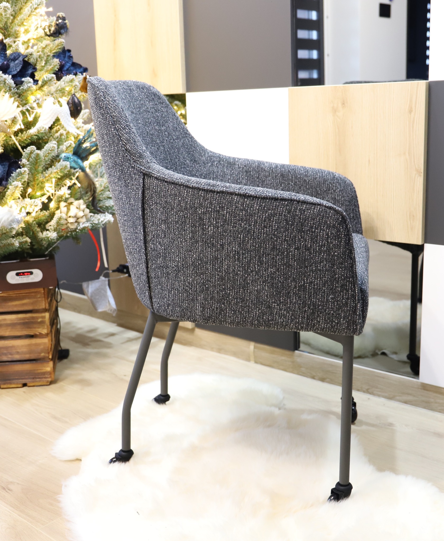 Szare tapicerowane krzesło z podłokietnikami na metalowych nogach z kółkami, ustawione na białym futrzanym dywaniku, obok choinki z niebieskimi ozdobami i drewnianej szafki z jasnymi frontami.