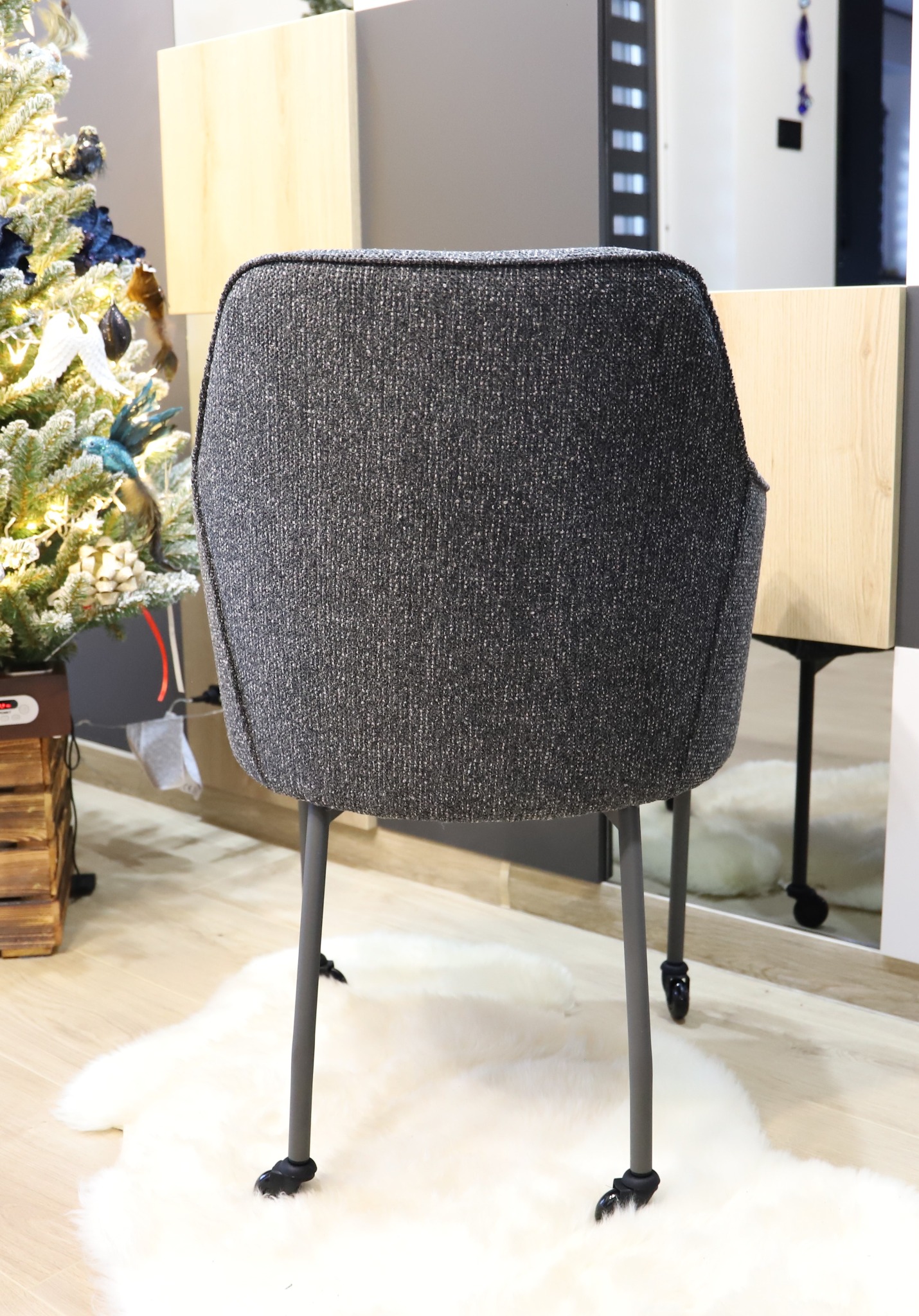 Tapicerowane krzesło z szarej tkaniny boucle na metalowych nogach z kółkami, na tle białego dywanu z długim włosiem i fragmentu choinki.