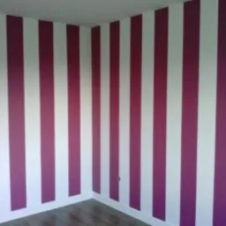 Wnętrze pokoju z białymi ścianami i pionowymi pasami w kolorze fuksji, widoczne drzwi po lewej stronie i fragment podłogi z jasnymi panelami.