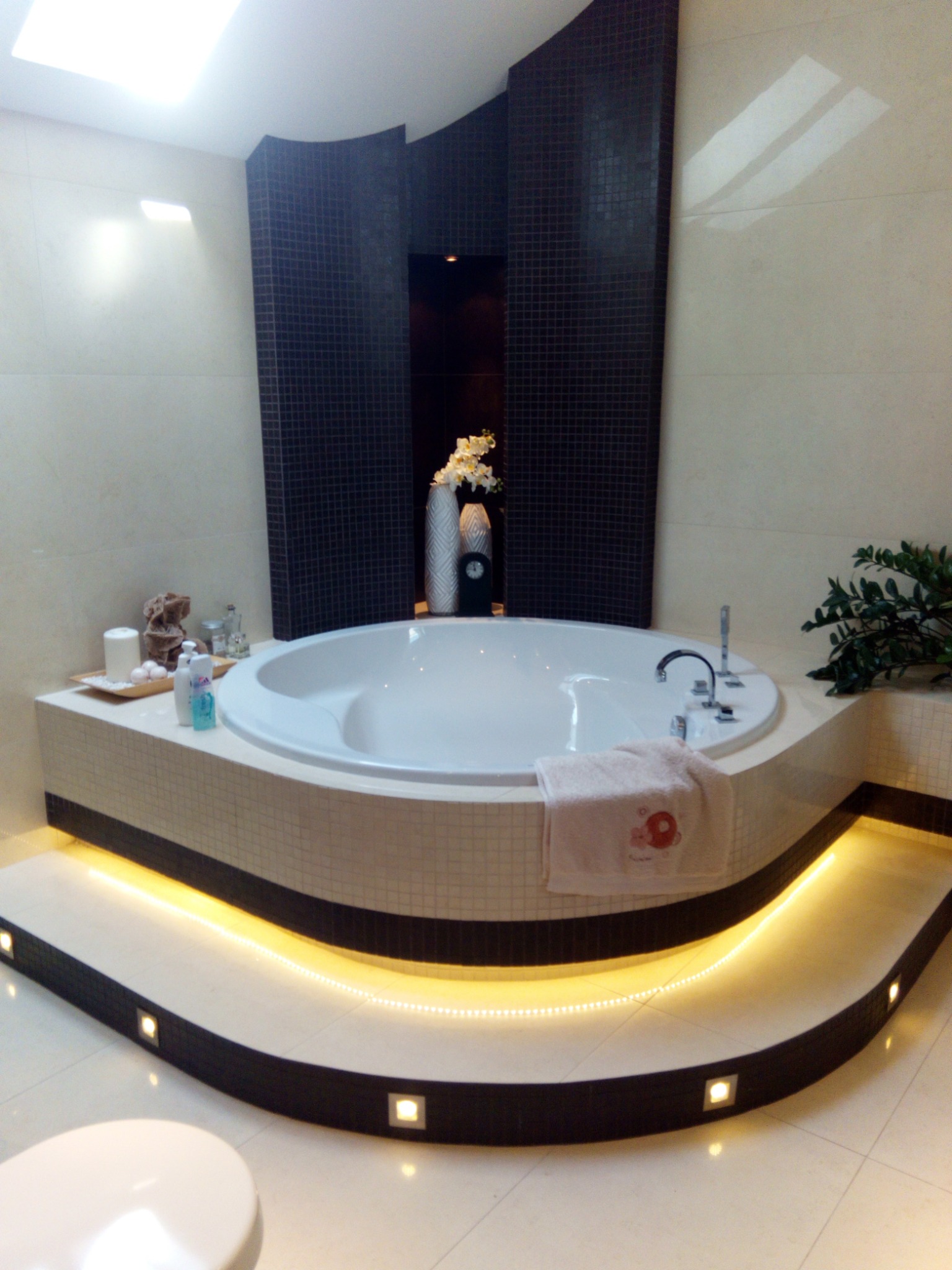 Nowoczesna łazienka z okrągłą wanną, podświetlanym stopniem, czarną mozaiką, beżowymi płytkami i dekoracjami na blacie.