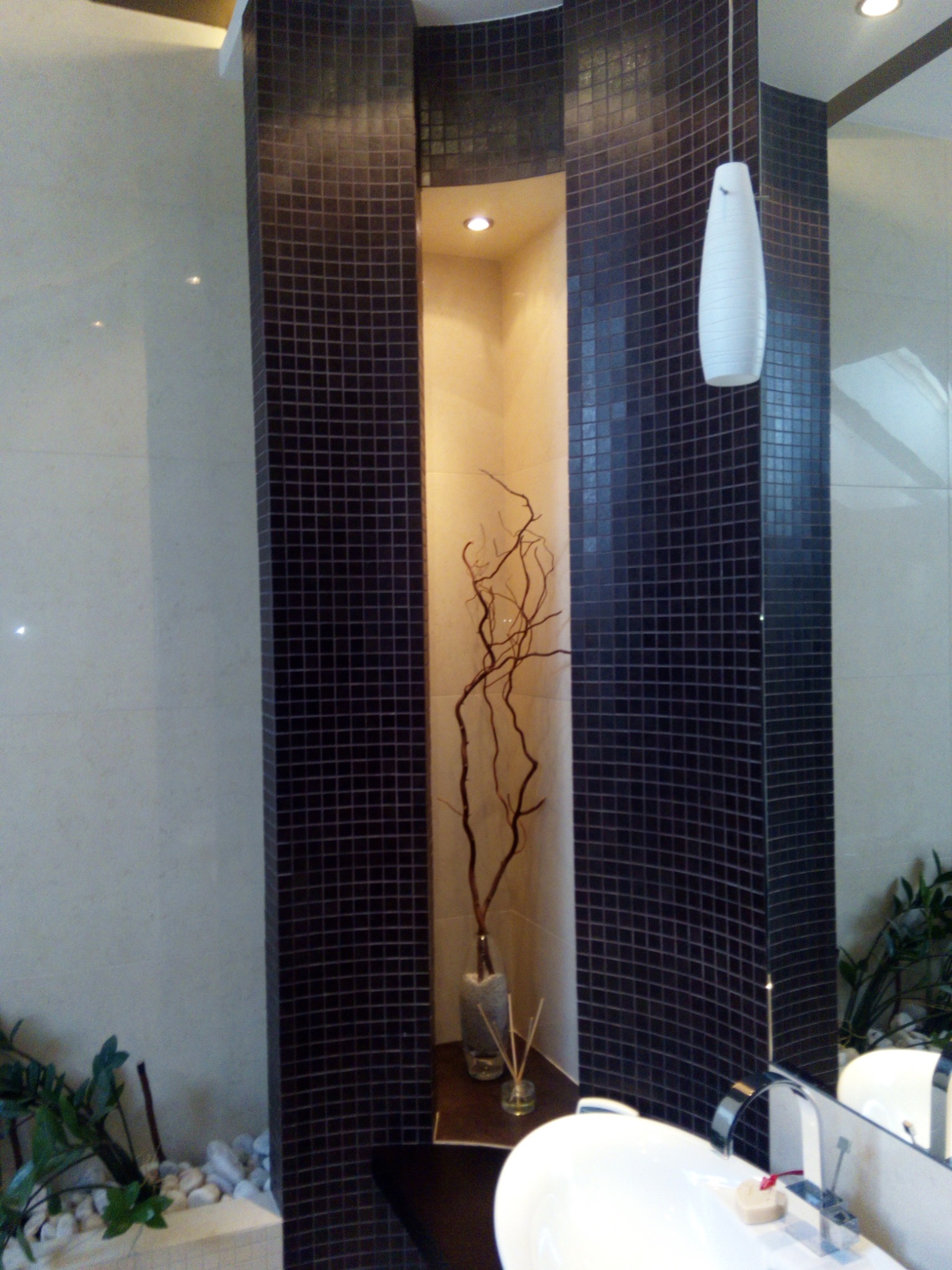 Nowoczesna łazienka z wnęką wyłożoną ciemną mozaiką, w której umieszczono dekoracyjne gałązki w szklanym wazonie, nad umywalką wisi biała, podłużna lampa.