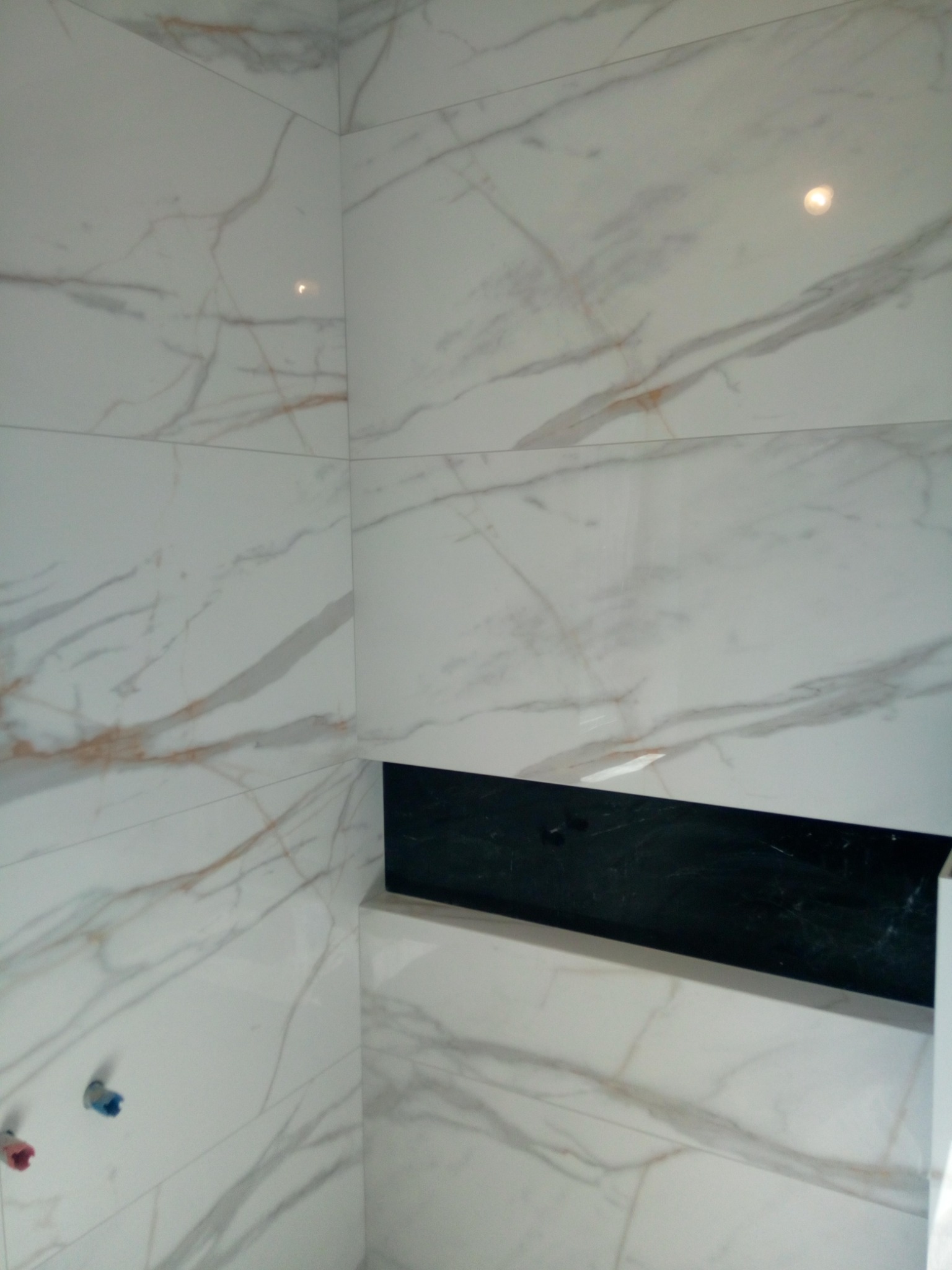 Ściana łazienki wyłożona białymi płytkami imitującymi marmur z widoczną wnęką z czarną okładziną i wystającymi końcówkami instalacji wodnej.