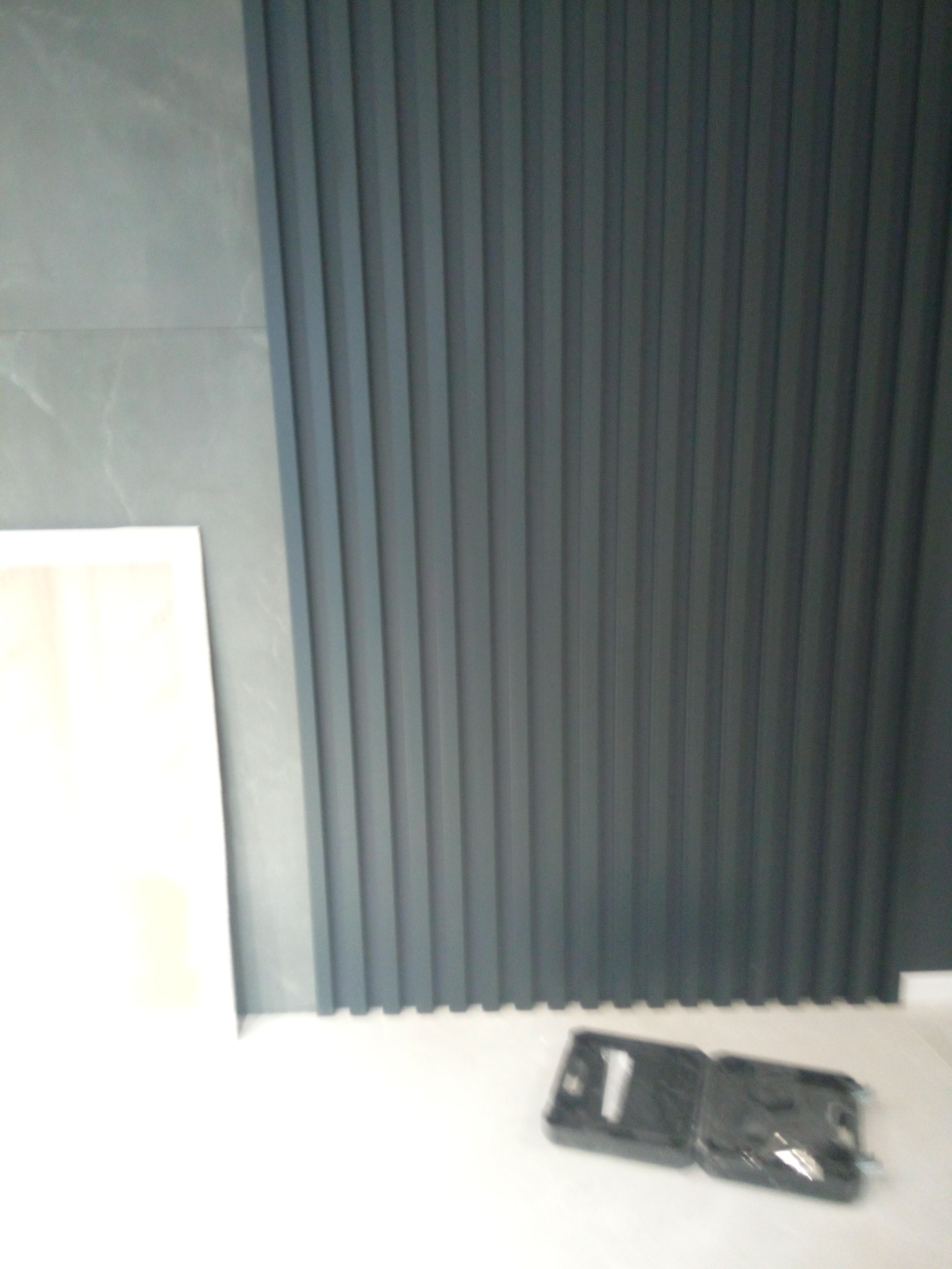 Szara ściana z pionowymi panelami dekoracyjnymi, obok płytka ścienna i rozłożona walizka narzędziowa na podłodze.