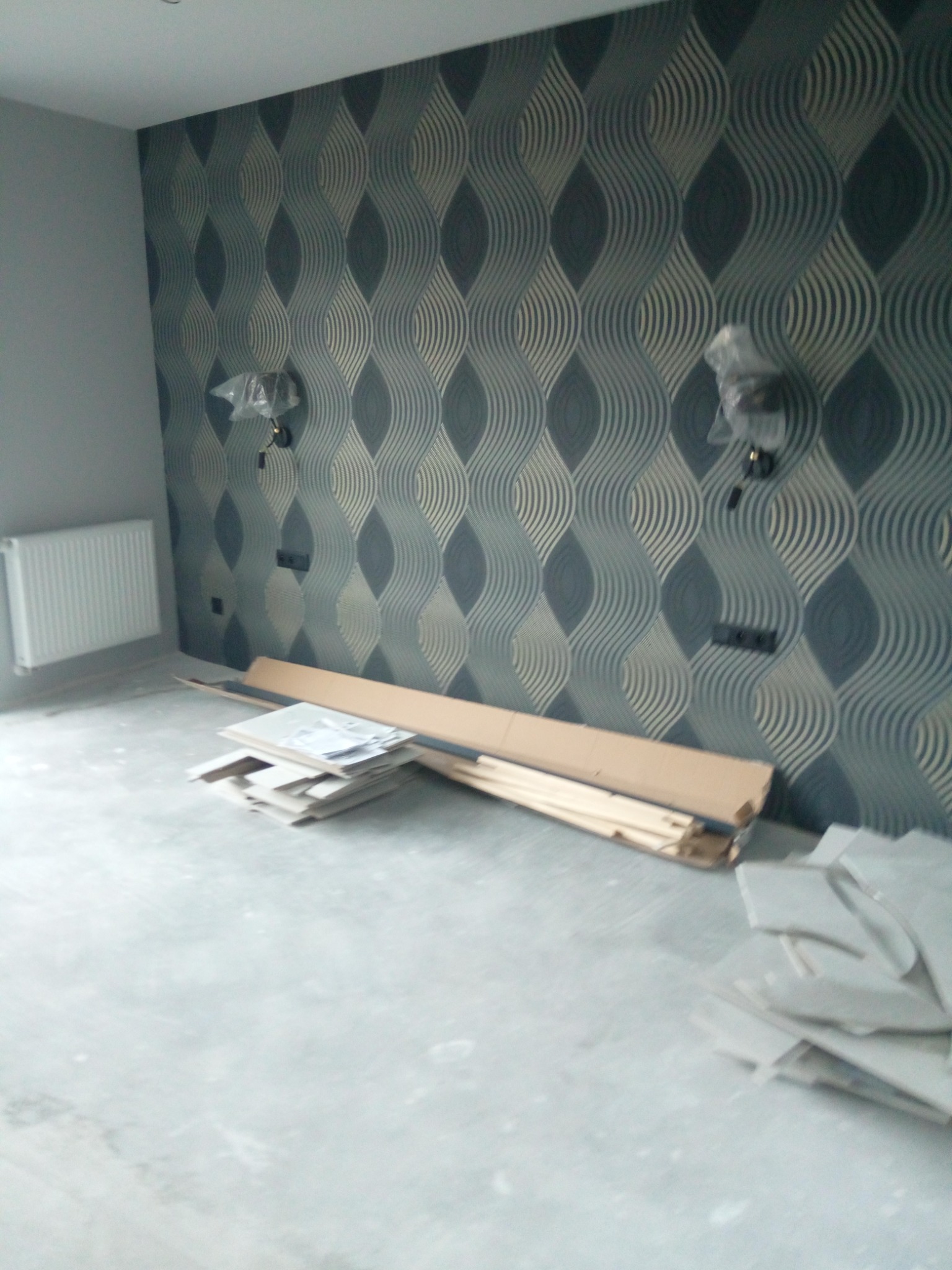 Ściana z tapetą o wzorze falistych linii w odcieniach szarości i beżu, zabezpieczone lampy ścienne folią, kartony i płytki na podłodze w trakcie prac wykończeniowych.