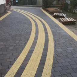 Ułożona kostka brukowa w odcieniach szarości z żółtymi, łukowatymi pasami. Na obrzeżach widoczne palety z kostką. Ujęcie z perspektywy.