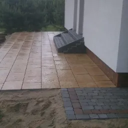 Ułożony taras z jasnobrązowej kostki brukowej, z kamiennymi schodami przy białym budynku, obok fragmentu ułożonej kostki granitowej w odcieniach szarości.