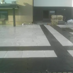 Nowoczesny taras z jasnoszarej kostki brukowej z czarnymi akcentami. Częściowo ułożona nawierzchnia, widoczne palety i materiały budowlane. Ujęcie z niskiej perspektywy.