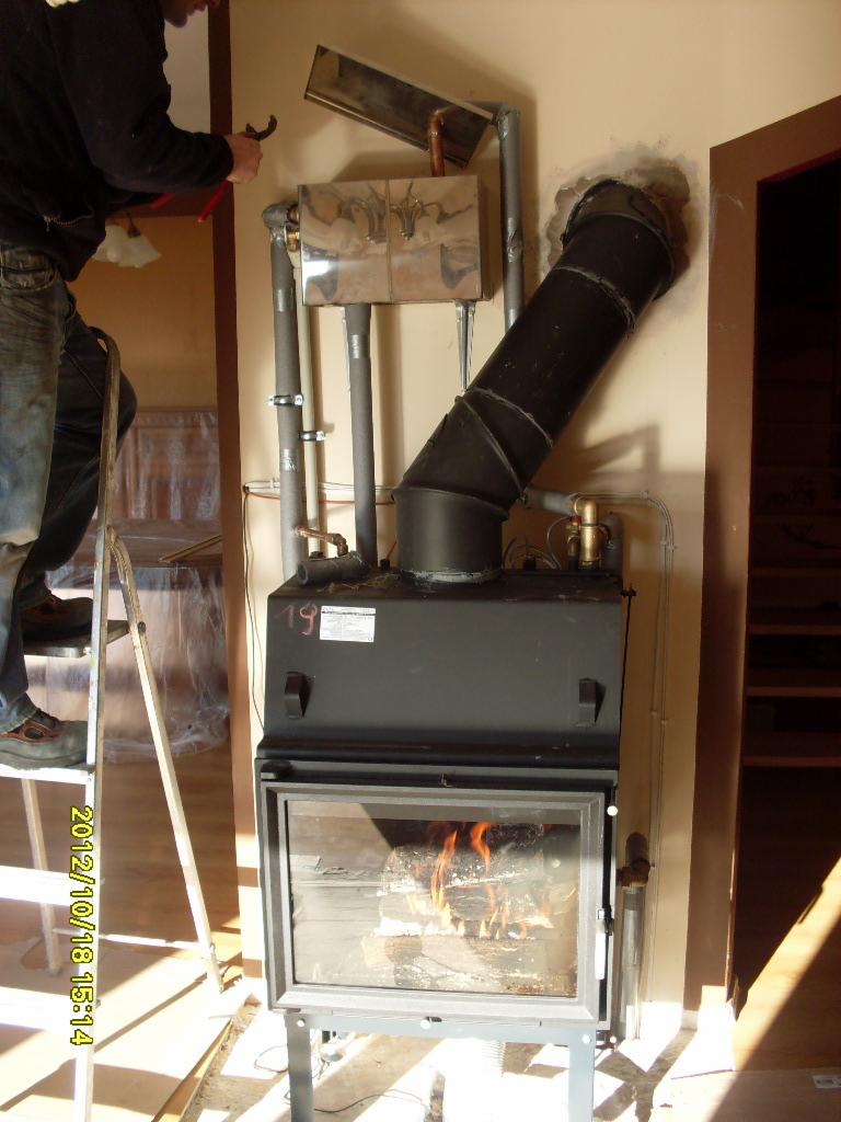 Instalacja kominka z otwartym paleniskiem, widoczne rury, kolanka i mocowania, hydraulik na drabinie używa klucza do rur.