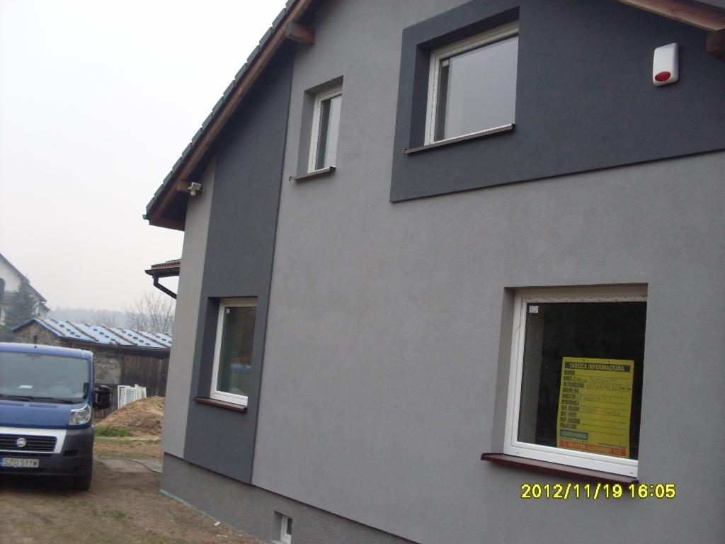 Elewacja domu po remoncie, z szarym tynkiem i ciemniejszymi szarymi akcentami wokół okien, na tle zaparkowany niebieski samochód dostawczy.