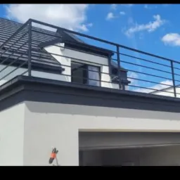 Nowoczesna, czarna balustrada metalowa na dachu budynku z białymi ścianami i szarą stolarką okienną na tle błękitnego nieba z chmurami.