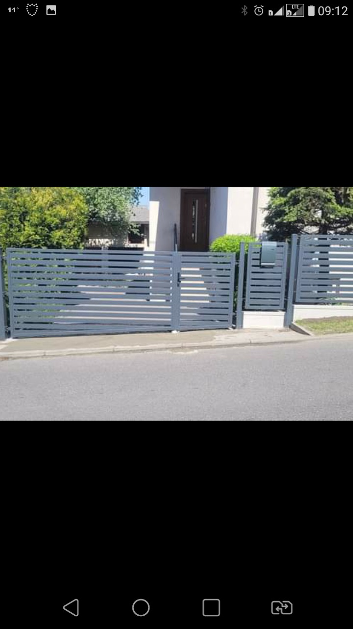 Nowoczesne, poziome ogrodzenie panelowe w kolorze szarym z furtką i bramą wjazdową, prowadzące do domu z ciemnymi drzwiami wejściowymi.