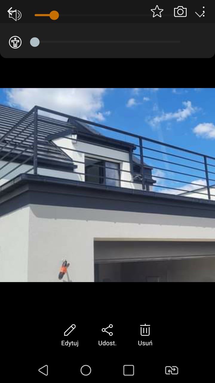 Nowoczesna, czarna balustrada metalowa na dachu budynku z białymi ścianami i szarą stolarką okienną na tle błękitnego nieba z chmurami.