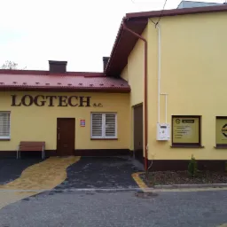 Żółty budynek firmy Logtech s.c. z brązowym dachem, widoczne okna z roletami, drzwi wejściowe i tabliczki informacyjne w oknach, przed budynkiem ławka i chodnik z kostki brukowej.