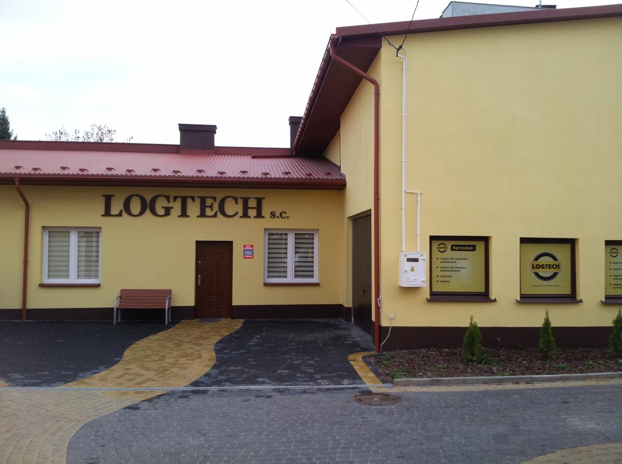 Żółty budynek firmy Logtech s.c. z brązowym dachem, widoczne okna z roletami, drzwi wejściowe i tabliczki informacyjne w oknach, przed budynkiem ławka i chodnik z kostki brukowej.