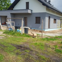 Styldom - Dwukondygnacyjny dom z szarą elewacją w trakcie prac wykończeniowych. Widoczne rusztowania i materiały budowlane na posesji. Dom z brązowymi oknami i czarnym dachem.