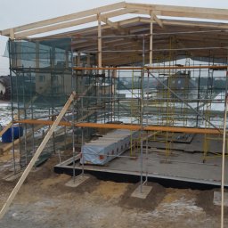 Styldom - Budowa domu szkieletowego w trakcie: widoczne drewniane elementy konstrukcyjne, rusztowania i fundamenty. Na pierwszym planie teren budowy z ziemią i resztkami śniegu.