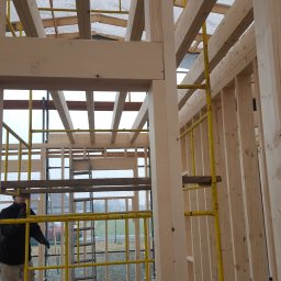 Styldom - Wnętrze domu szkieletowego w budowie, widoczne drewniane belki konstrukcyjne, żółte rusztowanie i częściowo widoczna postać pracownika w tle.