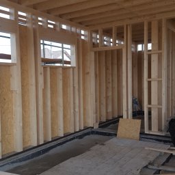 Styldom - Wnętrze domu w trakcie budowy szkieletowej: drewniana konstrukcja ścian, stropu i otwory okienne. Widoczne płyty OSB i fragment podłogi. Jasne, naturalne oświetlenie.