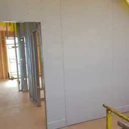 Wnętrze w trakcie montażu płyt karton-gips na metalowym stelażu. Widoczne drewniane elementy konstrukcji dachu i żółta folia zabezpieczająca.