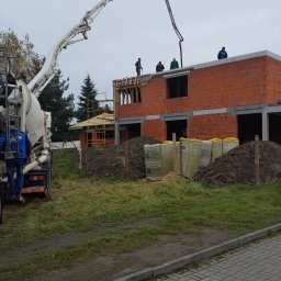 Styldom - Budowa domu z czerwonej cegły: betoniarka podaje beton na strop. Widoczni pracownicy na dachu i materiały budowlane na ziemi. Pochmurne niebo w tle.
