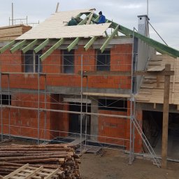 Styldom - Budowa domu z czerwonej cegły: ekipa montuje drewniane deski na zielonej konstrukcji dachu, widoczne rusztowania i stos drewna.