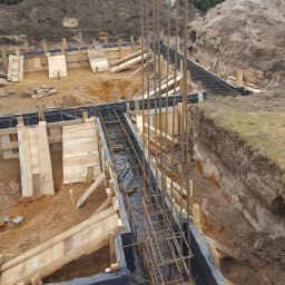 Styldom - Zbrojenie fundamentów domu jednorodzinnego w wykopie, zabezpieczone folią, z drewnianym szalunkiem. W tle ziemia i budynek.