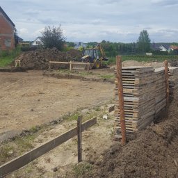 Styldom - Przygotowanie terenu pod budowę: wykopane fundamenty, drewniane szalunki, sterta płyt i koparka w tle. Prace ziemne w toku, widok na plac budowy.