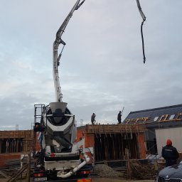 Styldom - Betonomieszarka z pompą na budowie domu z cegły. Widoczni robotnicy w trakcie pracy, szalunki i zbrojenia. Pochmurne niebo w tle.