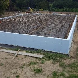 Zbrojony fundament płyty z izolacją pionową, widoczne zbrojenie z dystansami i folią. W tle pracownik z taczką. Drewniane stemple na ziemi.