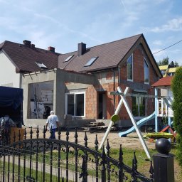 Styldom - Rozbudowa domu z czerwonej cegły, widok zza ozdobnego, czarnego ogrodzenia. Dodana część w stanie surowym, widoczne okna i dach. Plac zabaw w tle.