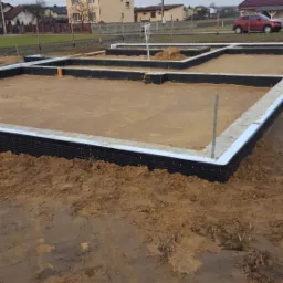 Przygotowany teren pod budowę domu: fundamenty z izolacją pionową, widoczny piasek i stalowe pręty, w tle zabudowa jednorodzinna i pochmurne niebo.