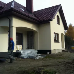 Jasny dom jednorodzinny z ciemną dachówką. Na pierwszym planie rusztowania i pracownik w niebieskiej kamizelce. Widoczne świeżo otynkowane ściany i detale architektoniczne.