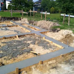 Zalane betonem szalunki fundamentów domu jednorodzinnego, widoczne zbrojenia w gruncie, ziemia i trawa w tle. Ujęcie z góry, w słoneczny dzień.