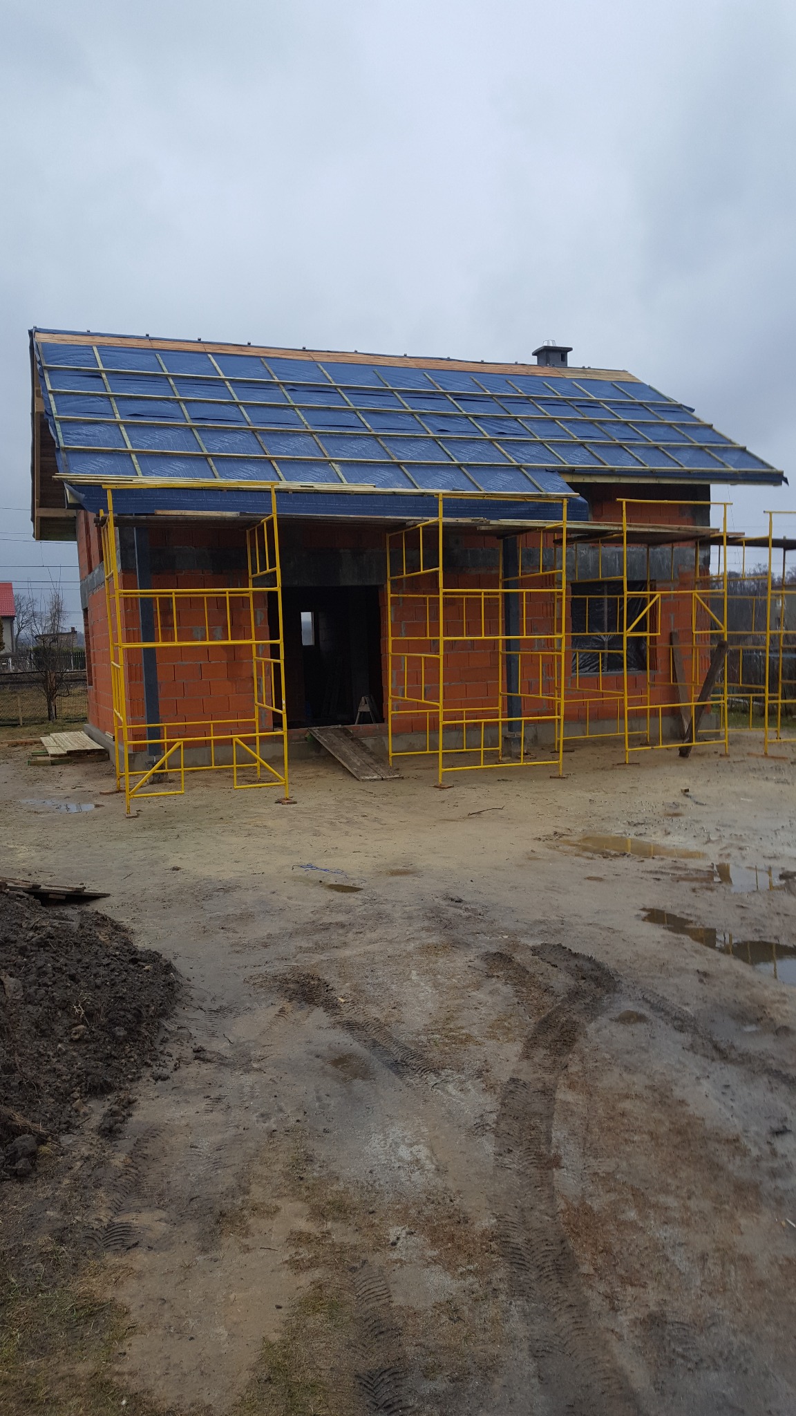 Budowa domu z czerwonej cegły z żółtym rusztowaniem i niebieską membraną dachową pod pochmurnym niebem. Widoczne ślady opon na błotnistym terenie.
