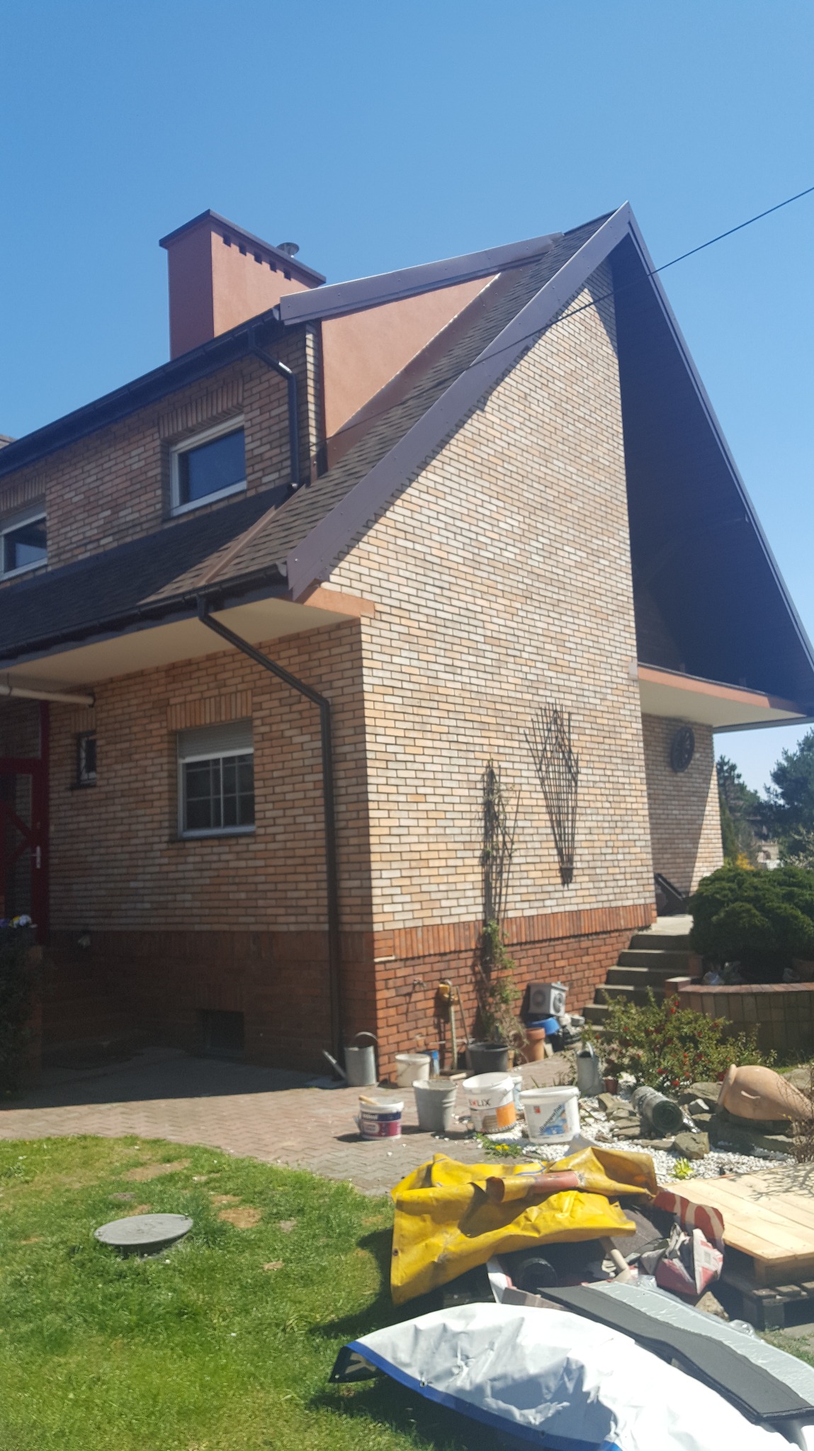 Dom z cegły klinkierowej po renowacji elewacji, widoczny fragment dachu, komin i teren wokół domu z materiałami budowlanymi.