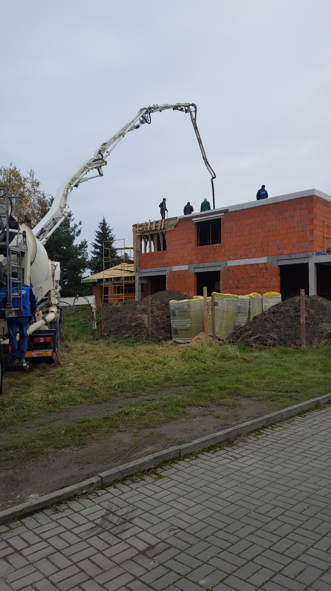 Budowa domu z czerwonej cegły: betoniarka podaje beton na strop. Widoczni pracownicy na dachu i materiały budowlane na ziemi. Pochmurne niebo w tle.