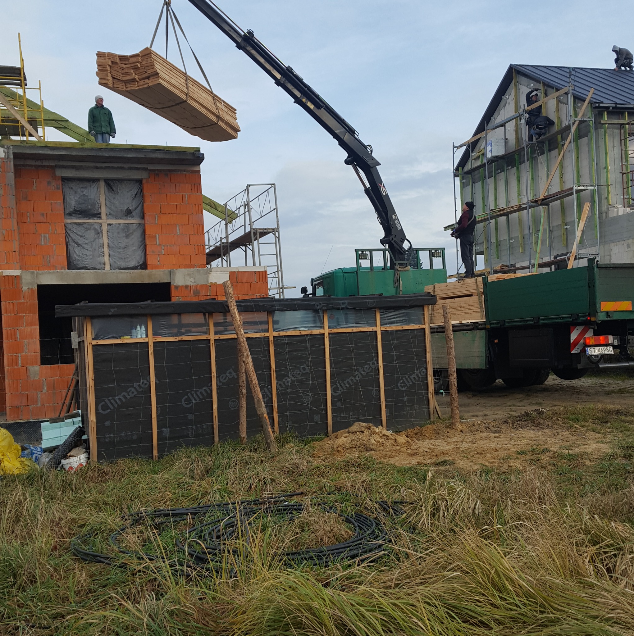 Budowa domu: dźwig podnosi pakiet desek na budynek z czerwonej cegły i szkieletowy. Na dachu pracownicy. Na pierwszym planie trawa i zwój czarnego kabla.