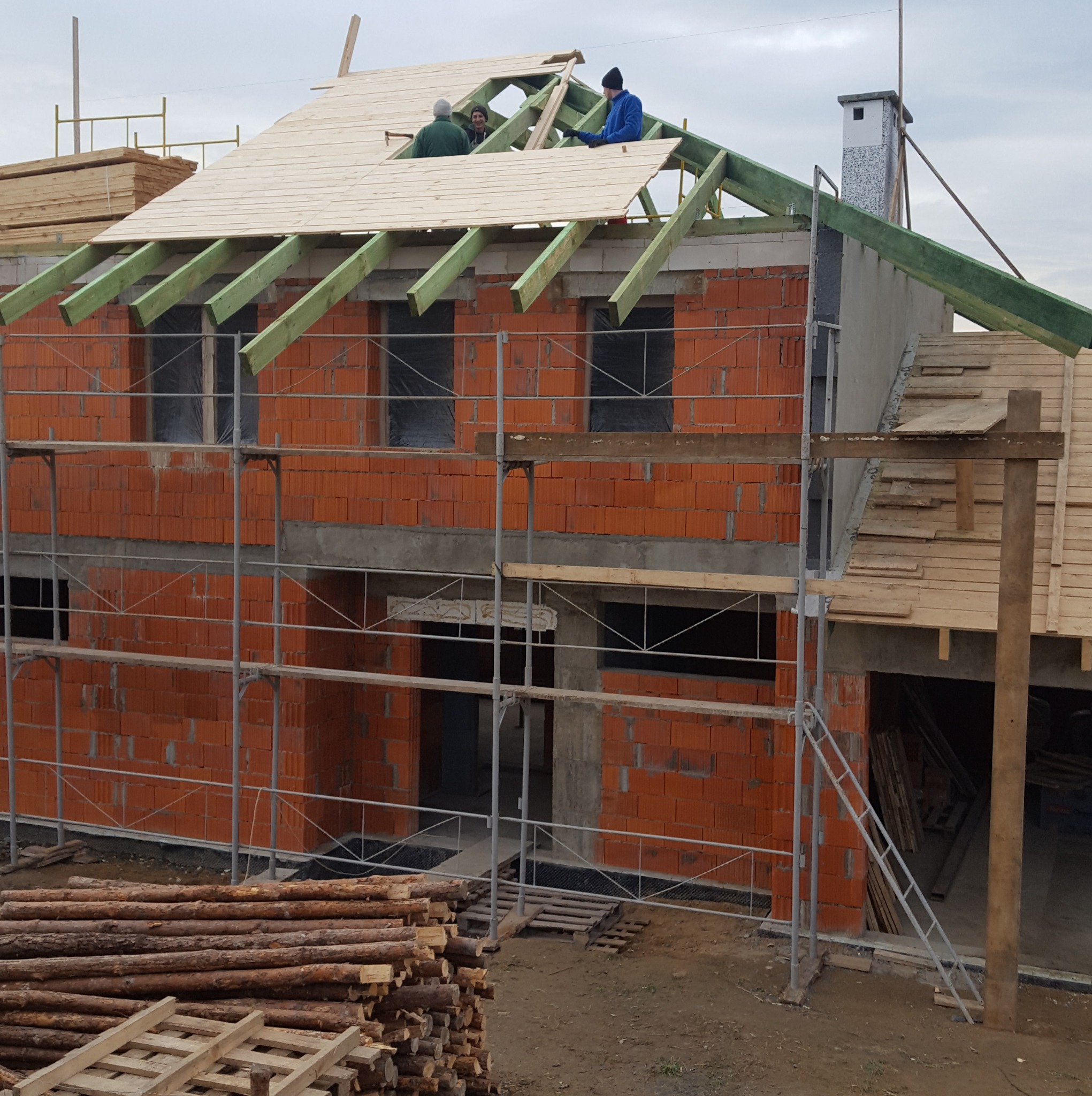 Budowa domu z czerwonej cegły: ekipa montuje drewniane deski na zielonej konstrukcji dachu, widoczne rusztowania i stos drewna.