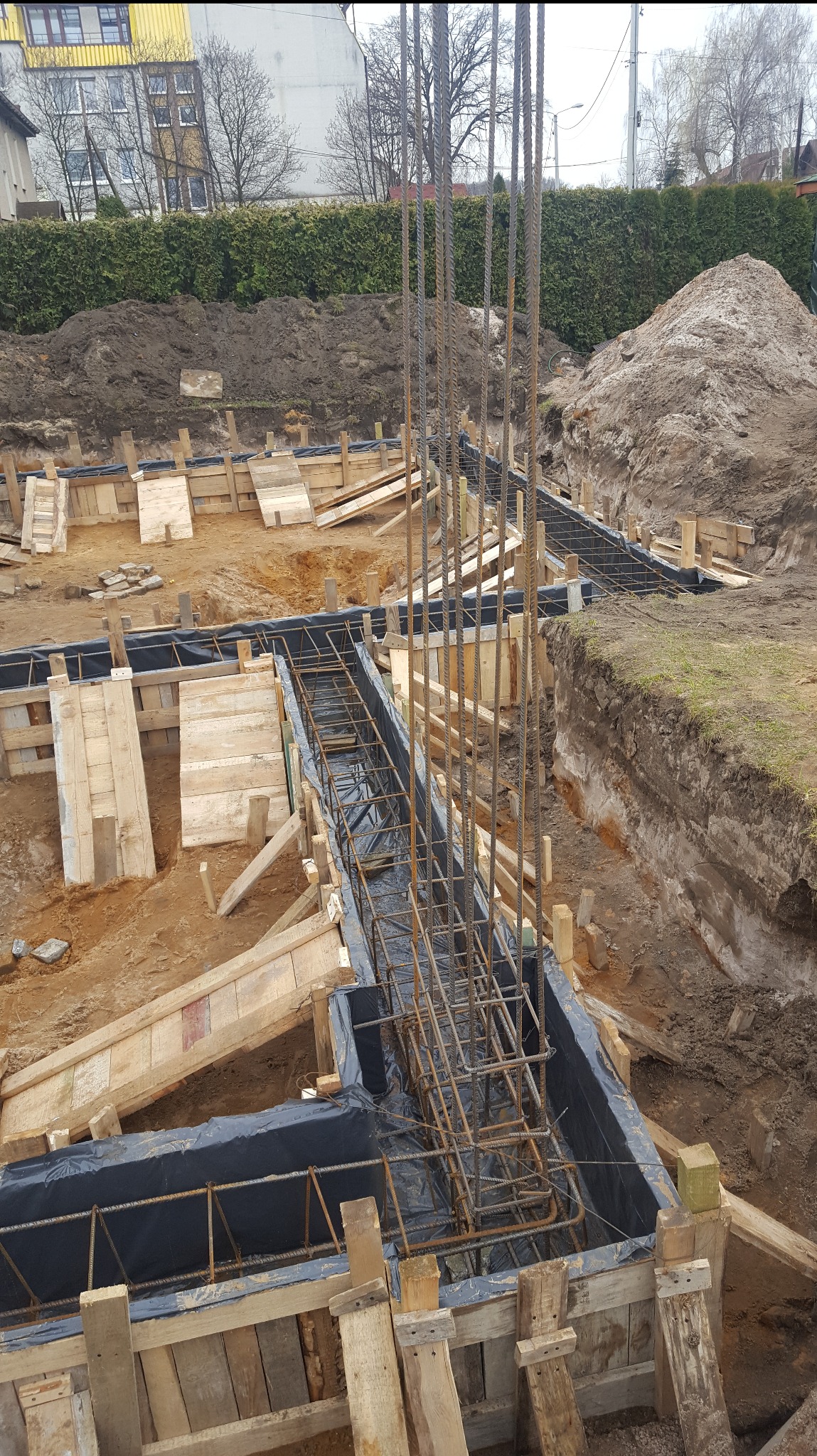 Zbrojenie fundamentów domu jednorodzinnego w wykopie, zabezpieczone folią, z drewnianym szalunkiem. W tle ziemia i budynek.