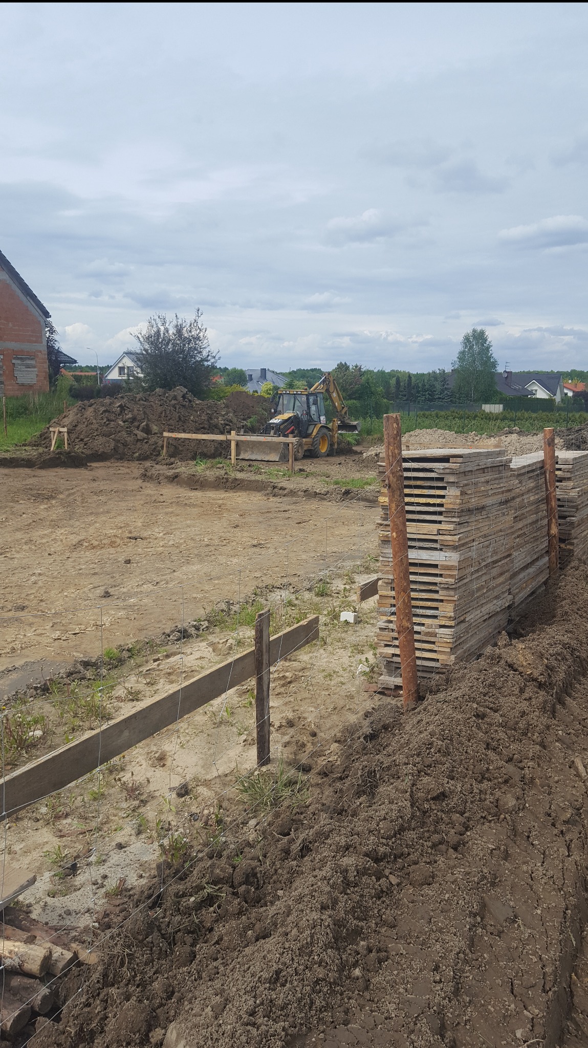Przygotowanie terenu pod budowę: wykopane fundamenty, drewniane szalunki, sterta płyt i koparka w tle. Prace ziemne w toku, widok na plac budowy.