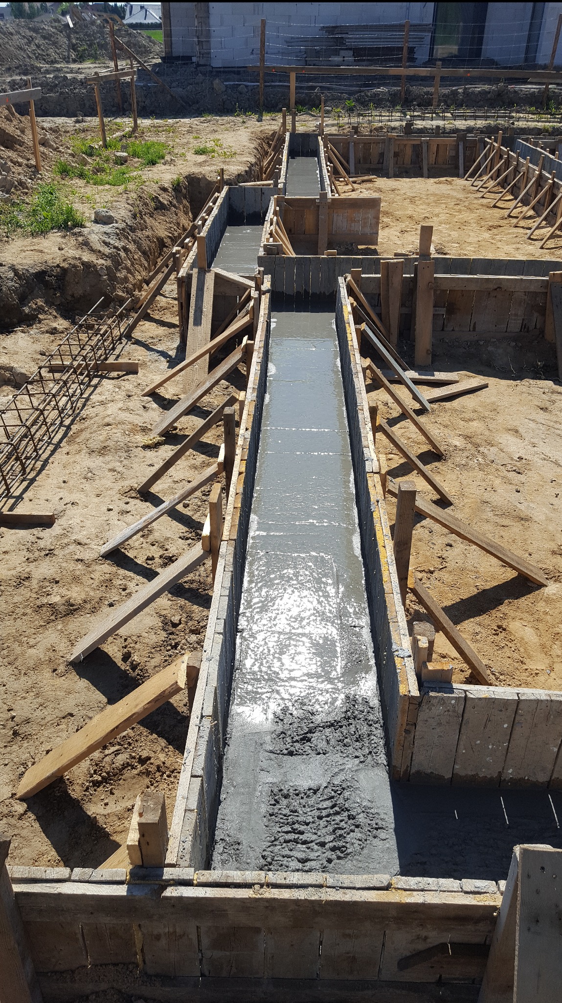 Zalewanie betonem szalunku fundamentów z widocznym zbrojeniem. Świeży beton wypełnia formę, przygotowaną do wylania, na piaszczystym podłożu.