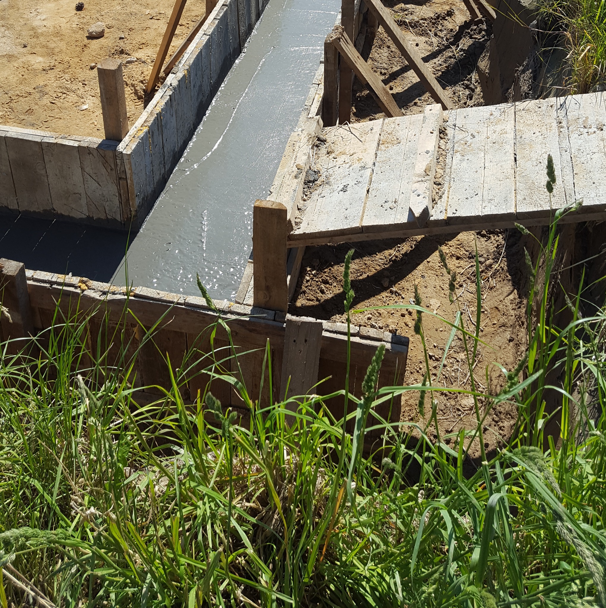 Zalewanie fundamentów w szalunku z desek. Widok z góry na świeży beton w wykopie, z zielenią w dolnej części kadru. Częściowo widoczny szalunek.