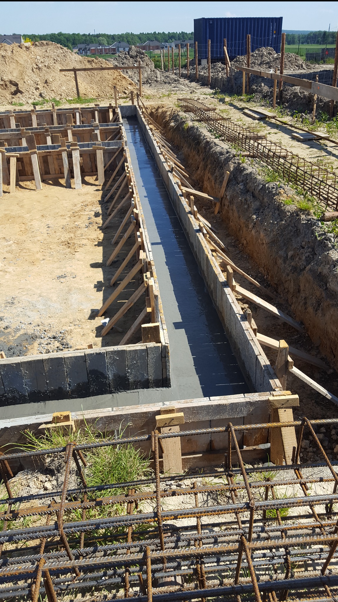 Zalewanie betonem szalunków fundamentowych pod budowę domu. Widoczne zbrojenia i drewniane konstrukcje. Ujęcie z góry, w słoneczny dzień.