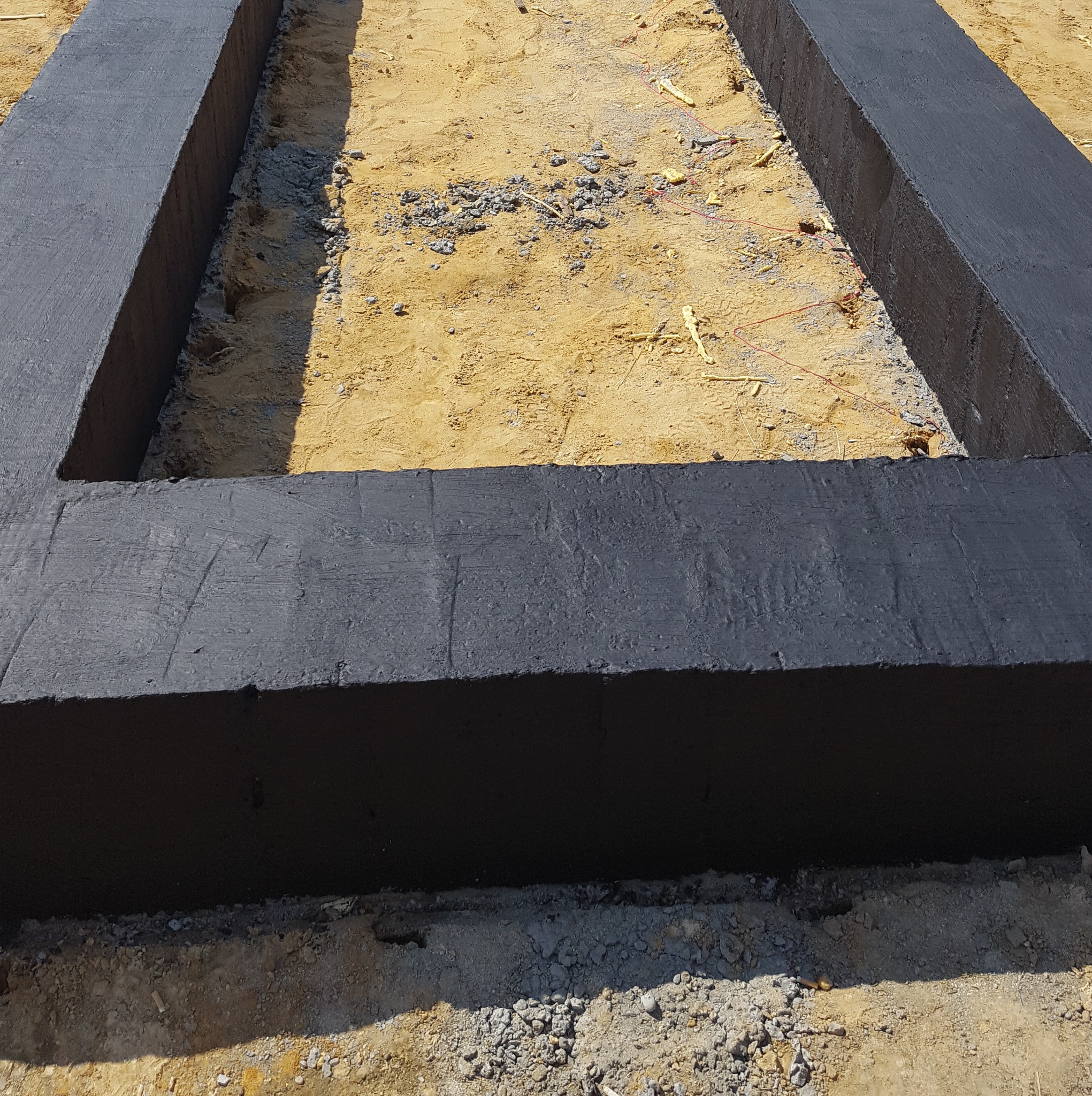 Zalane, pomalowane na czarno fundamenty pod budowę domu, widoczne z góry. W tle piasek i wystające elementy zbrojenia.